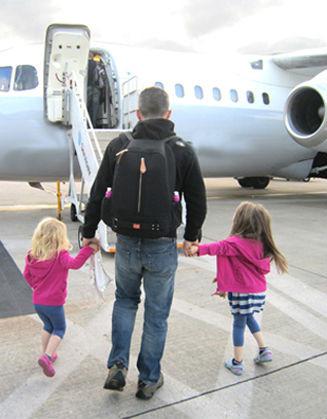 Man walking towards a plane with the backpack on his back while holding the hands of two young children